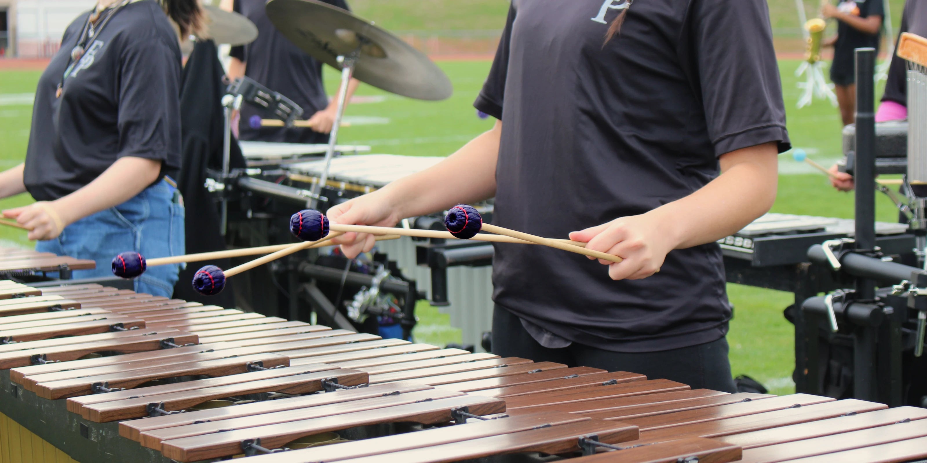 Person playing a marimba on a sports field with other band members in the background.