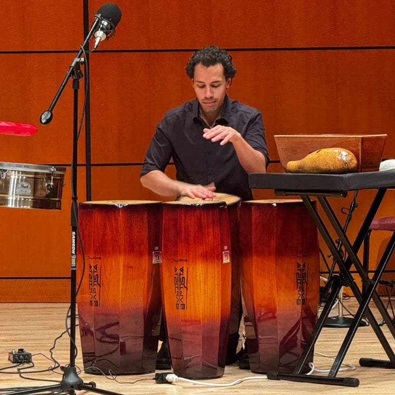 Man playing conga drums in a studio setting with musical equipment around.