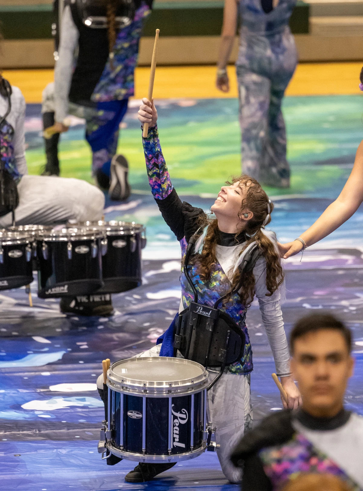 Person playing a drum with raised arm on a stage with other performers.