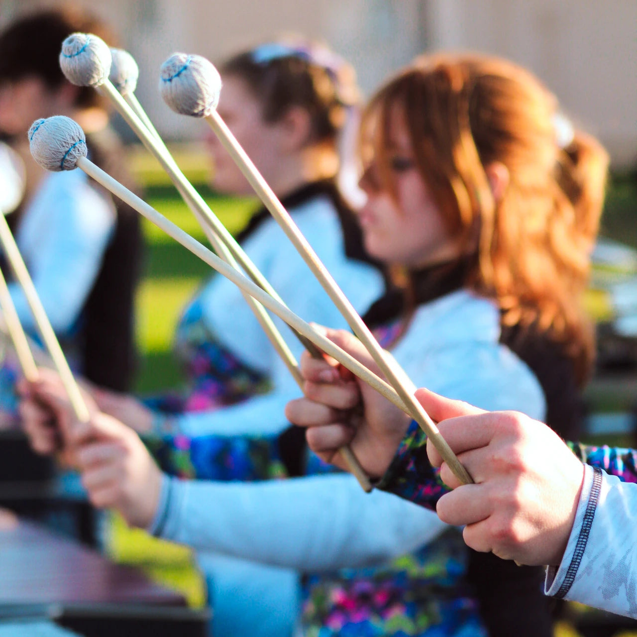 Close-up of hands holding mallets with a blurred background of people playing instruments.