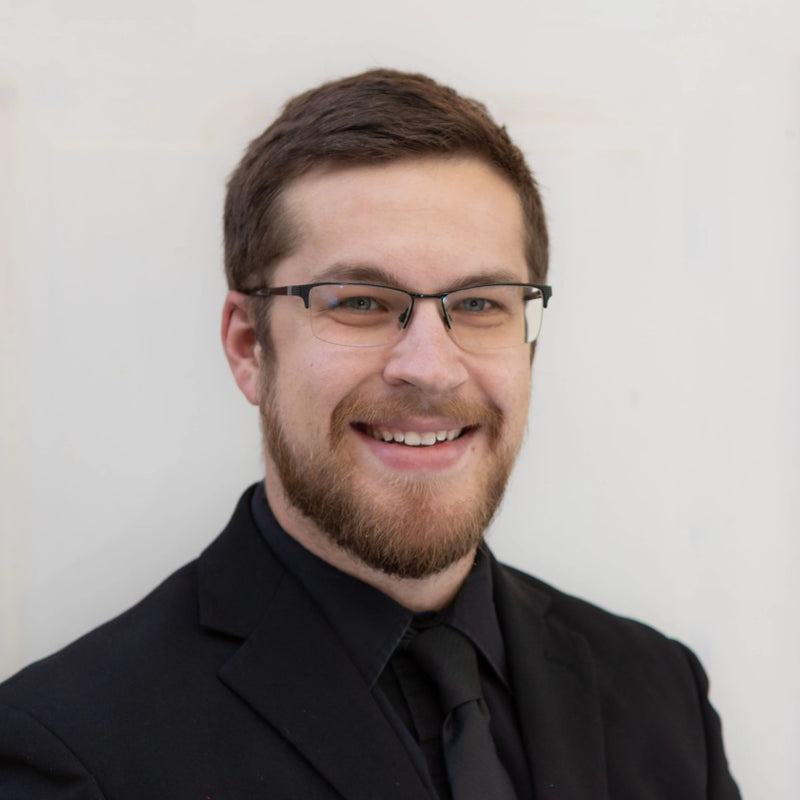 Man wearing glasses and a black suit against a plain background